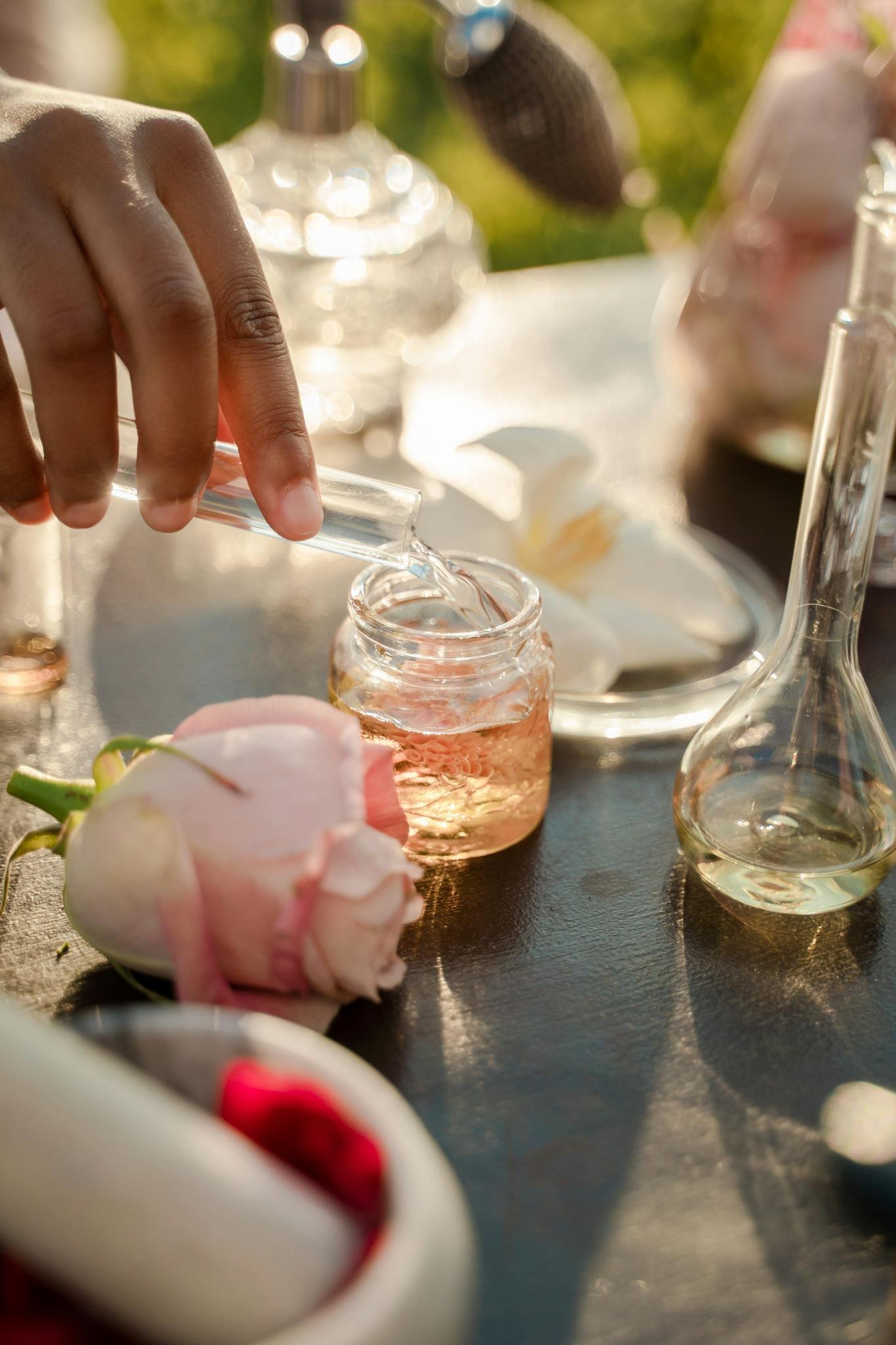 Hand pouring floral essence into a small glass jar, surrounded by roses and perfume bottles.